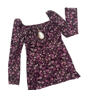 May include: A dark purple dress with a floral pattern, long sleeves, and a square neckline. The dress has a ruffled hem and a pendant necklace. The floral pattern includes pink and white flowers and paisley shapes.