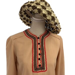 May include: A tan-colored blouse with a decorative red, green, and black trim around the neckline and a row of black buttons down the front. A crocheted sun hat in brown and beige sits atop a mannequin head.