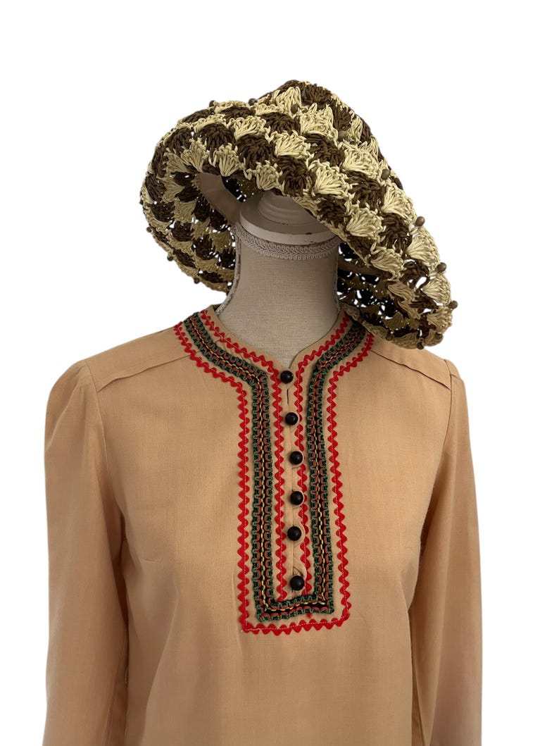 May include: A tan-colored blouse with a decorative red, green, and black trim around the neckline and a row of black buttons down the front. A crocheted sun hat in brown and beige sits atop a mannequin head.