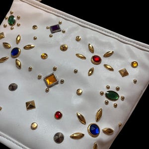 May include: White clutch bag adorned with colorful gemstones and gold-toned embellishments. The bag features a variety of jewel-toned accents, including green, blue, red, and yellow, along with small gold studs and leaf-shaped details.