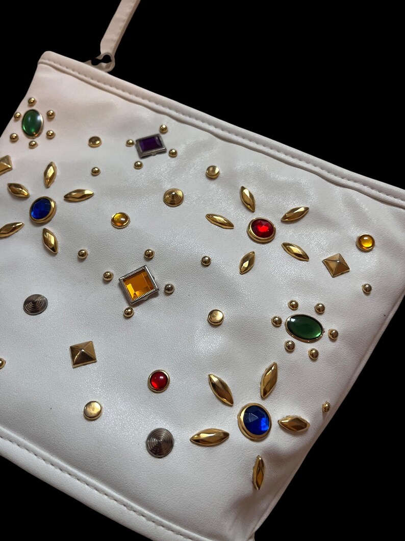 May include: White clutch bag adorned with colorful gemstones and gold-toned embellishments. The bag features a variety of jewel-toned accents, including green, blue, red, and yellow, along with small gold studs and leaf-shaped details.