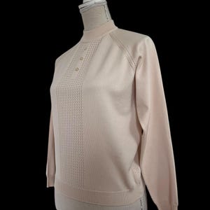 May include: A pale pink long-sleeved jumper with a mock neck and three buttons. The jumper features a textured design down the front and is displayed on a mannequin against a black backdrop.