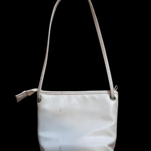 May include: A cream-colored shoulder bag with a rectangular body and a thin strap. The bag has a simple design with a small bow detail near the strap attachment. The background is solid black.