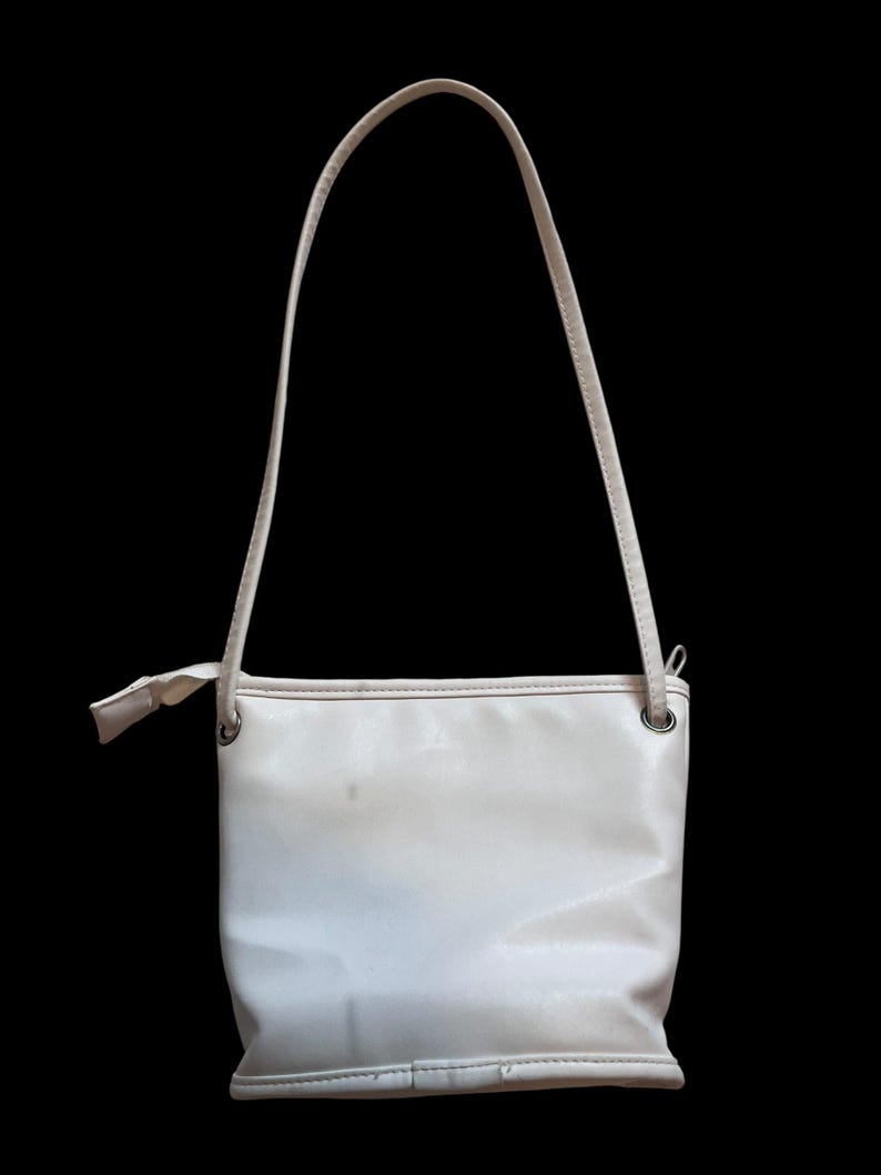 May include: A cream-colored shoulder bag with a rectangular body and a thin strap. The bag has a simple design with a small bow detail near the strap attachment. The background is solid black.