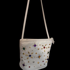May include: White shoulder bag adorned with colorful gemstones and gold-tone studs. The bag features a long, thin strap and a rounded bottom. The gemstones are various shapes and colors, including red, purple, green, and yellow.