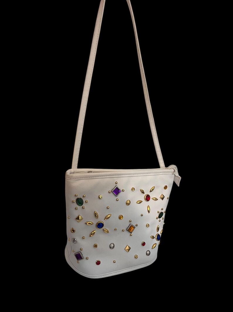 May include: White shoulder bag adorned with colorful gemstones and gold-tone studs. The bag features a long, thin strap and a rounded bottom. The gemstones are various shapes and colors, including red, purple, green, and yellow.