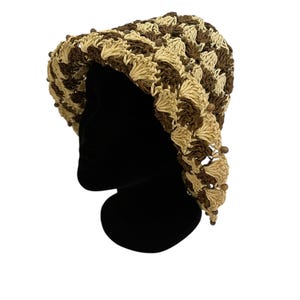 May include: A crocheted hat in shades of brown and beige, displayed on a black mannequin head. The hat features a textured pattern and is adorned with small, dark brown beads along the brim. The hat is a stylish accessory.