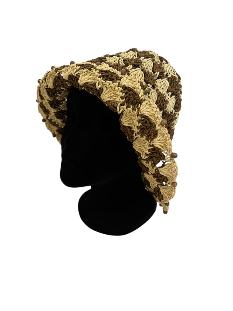 May include: A crocheted hat in shades of brown and beige, displayed on a black mannequin head. The hat features a textured pattern and is adorned with small, dark brown beads along the brim. The hat is a stylish accessory.