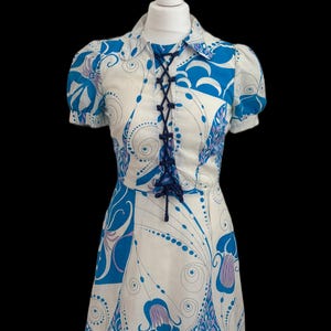 May include: A vintage dress displayed on a mannequin. The dress has a white base with a blue and purple floral design. It features short sleeves, a collared neckline, and a lace-up front. The dress is knee-length.