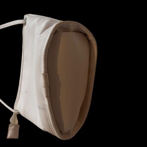 May include: A light beige shoulder bag with a long, thin strap and a small, square accent piece. The bag has a rounded shape and a subtle sheen, set against a black background.