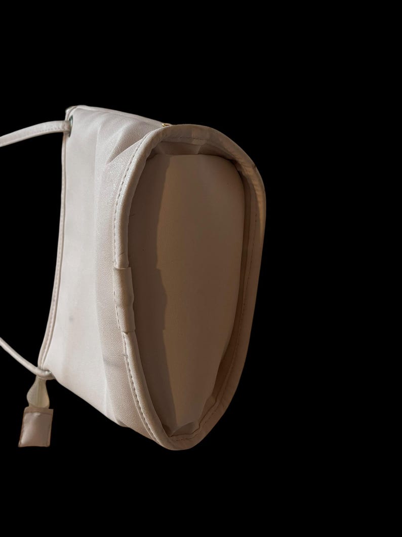 May include: A light beige shoulder bag with a long, thin strap and a small, square accent piece. The bag has a rounded shape and a subtle sheen, set against a black background.