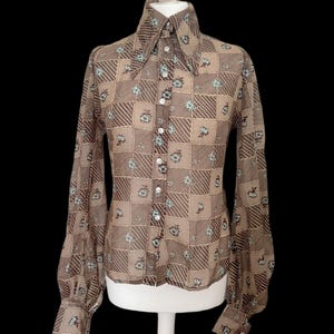 May include: A long-sleeved button-down shirt featuring a brown and tan checkered pattern with small light blue floral accents. The shirt has a large pointed collar, full sleeves with buttoned cuffs, and a front placket with white buttons.