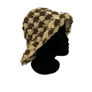 May include: A crocheted bucket hat in a checkerboard pattern of brown and beige. The hat has a wide brim and is displayed on a black mannequin head. The hat is made of a textured yarn.