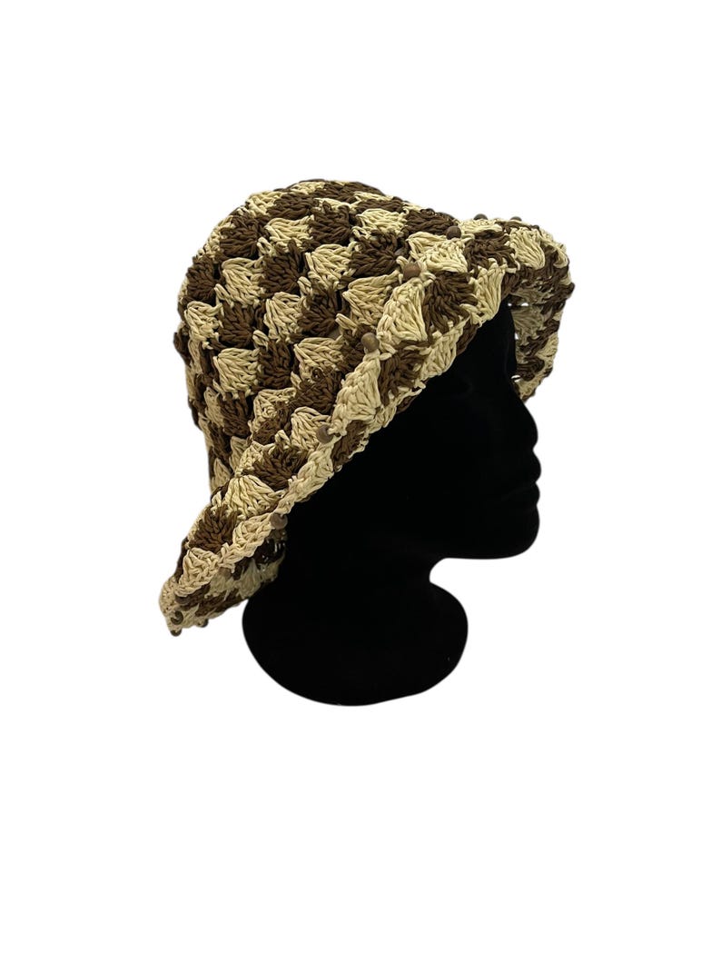 May include: A crocheted bucket hat in a checkerboard pattern of brown and beige. The hat has a wide brim and is displayed on a black mannequin head. The hat is made of a textured yarn.