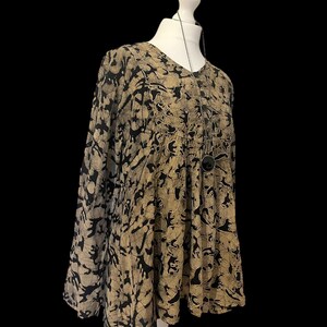 May include: A beige and black patterned top with long sleeves and a V-neck. The top is displayed on a white mannequin and features a button-down front. A silver necklace with a circular pendant is also visible. The fabric has a loose, flowing fit.