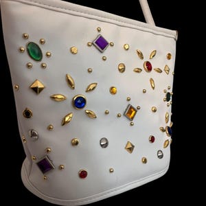 May include: White handbag adorned with colorful gemstones and gold-toned studs. The bag features a shoulder strap and a rounded base. The gemstones are in various shapes and colors, including green, purple, red, and blue.
