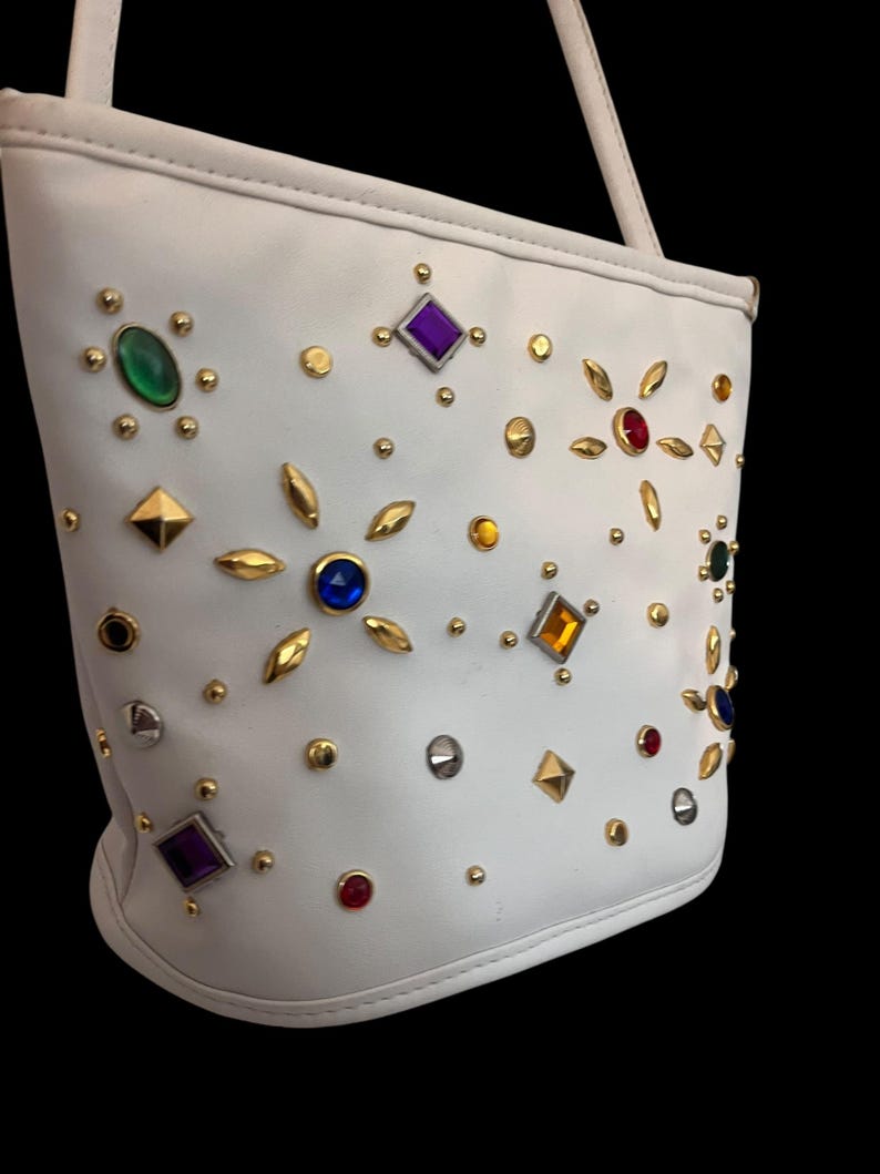 May include: White handbag adorned with colorful gemstones and gold-toned studs. The bag features a shoulder strap and a rounded base. The gemstones are in various shapes and colors, including green, purple, red, and blue.