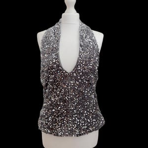May include: A sleeveless halter neck top covered in small, reflective silver sequins. The garment features a deep, plunging V-neckline and a fitted silhouette, displayed on a white mannequin against a solid black background.