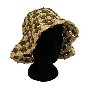 May include: A crocheted sun hat in shades of brown and beige. The hat features a wide brim and is adorned with small wooden beads around the edge. The hat is displayed on a black mannequin head against a white background.
