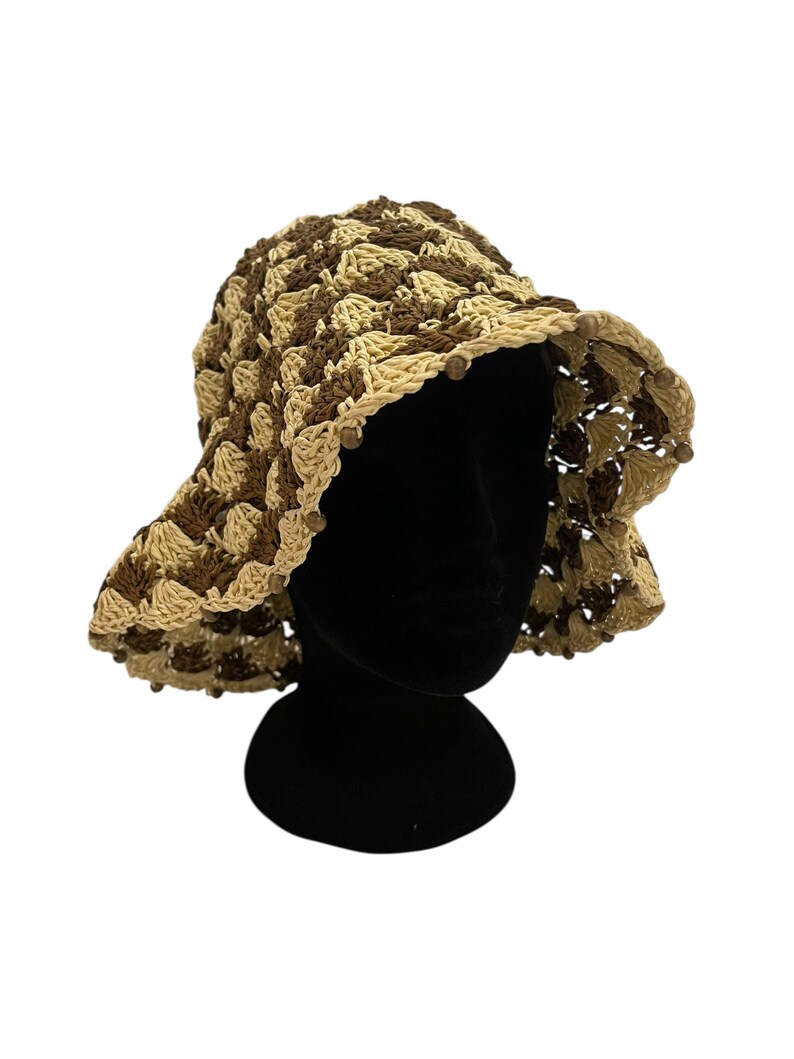 May include: A crocheted sun hat in shades of brown and beige. The hat features a wide brim and is adorned with small wooden beads around the edge. The hat is displayed on a black mannequin head against a white background.