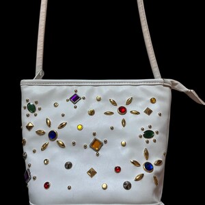 May include: White crossbody bag adorned with colorful gemstones and gold-toned studs. The bag features a long, thin strap and a rectangular shape. The gemstones are in various shapes and colors, including red, blue, green, and purple.