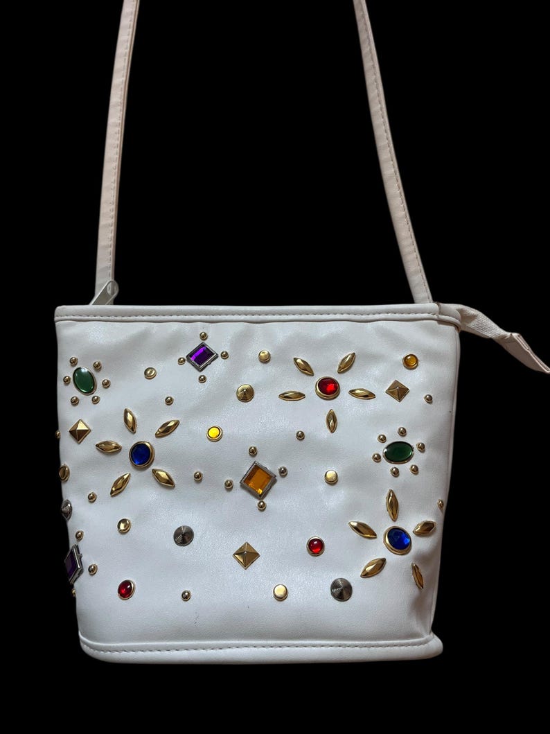 May include: White crossbody bag adorned with colorful gemstones and gold-toned studs. The bag features a long, thin strap and a rectangular shape. The gemstones are in various shapes and colors, including red, blue, green, and purple.
