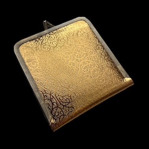 May include: A gold-coloured, rectangular compact with an ornate, swirling pattern. The compact has a silver-coloured frame and a small handle. The background is black.