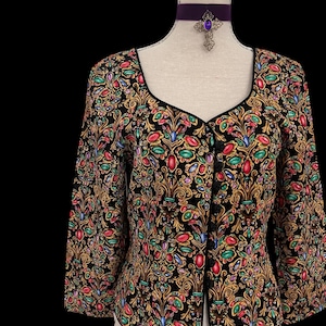May include: A black, long-sleeved jacket with a jewel-toned floral pattern. The jacket features a black trim and a scoop neckline. A purple velvet choker with a cross pendant is also visible.
