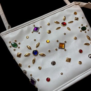 May include: White crossbody bag adorned with colorful gemstones and gold-toned studs. The bag features a long strap and is decorated with various shapes and colors of gems, including red, blue, green, and purple.
