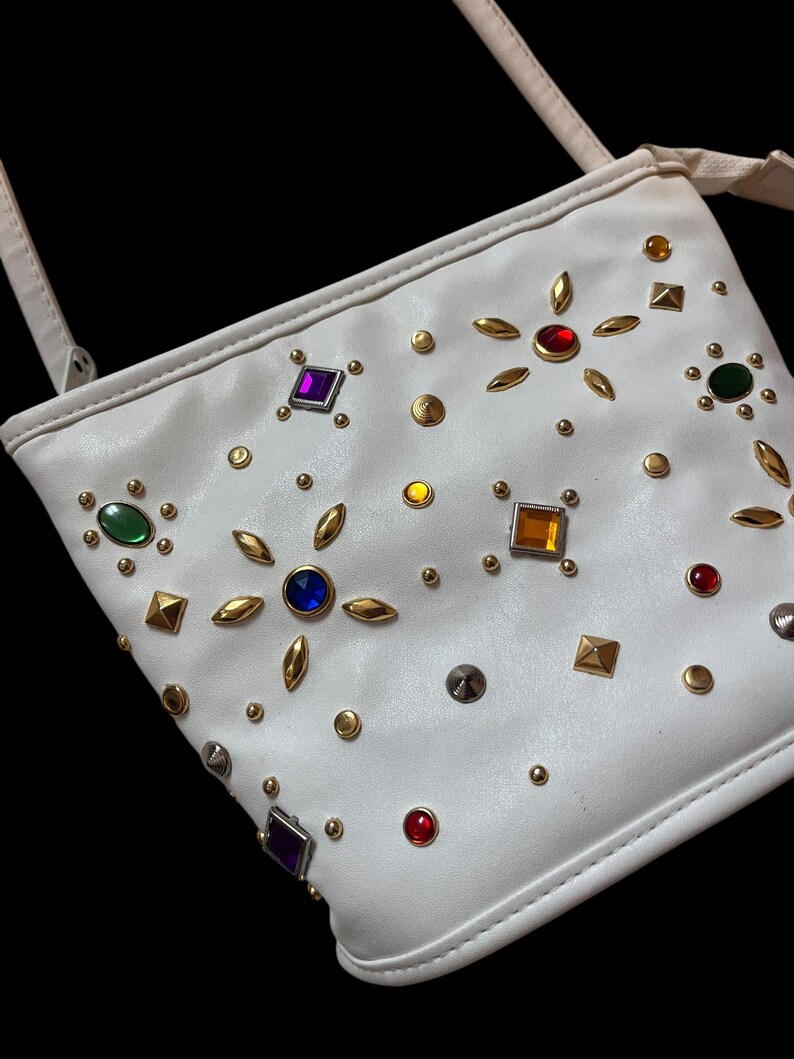 May include: White crossbody bag adorned with colorful gemstones and gold-toned studs. The bag features a long strap and is decorated with various shapes and colors of gems, including red, blue, green, and purple.