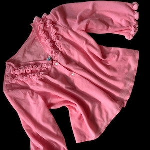May include: A pink, long-sleeved blouse with ruffled trim around the neckline and cuffs. The blouse has a single button closure at the neckline.