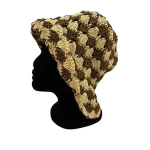 May include: A crocheted hat in a checkerboard pattern of light beige and brown. The hat is displayed on a black mannequin head against a white background. The hat's texture is visible, showcasing the handmade quality.