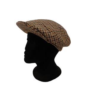 May include: A brown and tan plaid flat cap. The cap is made of a woven fabric with a classic houndstooth pattern. The cap is displayed on a black mannequin head.