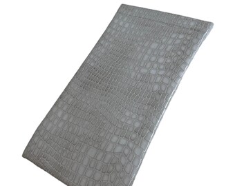 May include: A rectangular, light gray pouch with a textured, crocodile-like pattern. The pouch is made of a shiny material and appears to be empty. The pouch is angled slightly, showing its shape and texture against a white background.