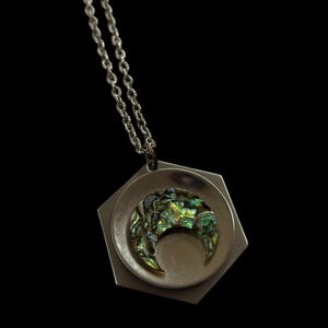 May include: Silver necklace with a hexagonal pendant. The pendant has a circular inset with a crescent moon design made of iridescent abalone shell. The necklace chain is a delicate, twisted link style, and the background is black.