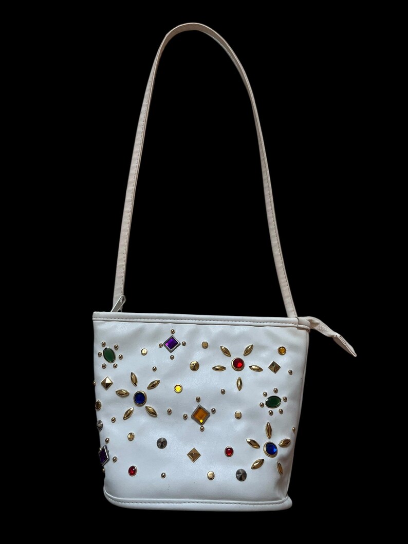 May include: White handbag with a single strap, adorned with colorful gemstones and gold-tone studs. The bag has a cylindrical shape and a cream-colored strap. The gemstones are in various shapes and colors, including red, blue, green, and yellow.