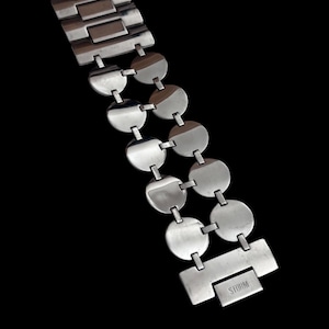 May include: A silver-toned metal watch bracelet featuring a series of circular links arranged in two parallel rows. The bracelet includes a rectangular clasp engraved with the word STORM, connected to a wider, flat-link section at the top end.