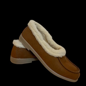 May include: A pair of brown moccasin-style slippers with a cream-coloured, fluffy lining. The slippers have a rounded toe and a light brown sole. The background is black.