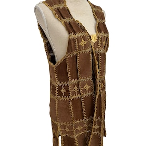 May include: Brown suede vest with a patchwork design. The vest has a V-neck and a gold-coloured sunburst clasp. The edges are trimmed with gold stitching. The vest is displayed on a mannequin.