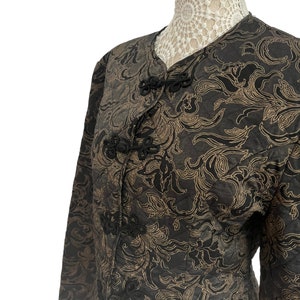 May include: A black and gold brocade jacket with long sleeves and a mandarin collar. The jacket has black knotted closures and a floral pattern.