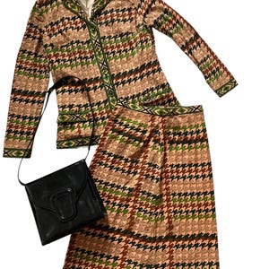 May include: A two-piece outfit featuring a long-sleeved jacket and a skirt. Both pieces are made from a brown, green and pink fabric with a houndstooth pattern. The jacket has a collar and a button fastening. The skirt is knee-length and has a waistband. A black leather handbag is included.