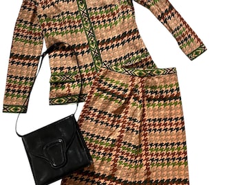 May include: A two-piece outfit featuring a long-sleeved jacket and a skirt. Both pieces are made from a brown, green and pink fabric with a houndstooth pattern. The jacket has a collar and a button fastening. The skirt is knee-length and has a waistband. A black leather handbag is included.