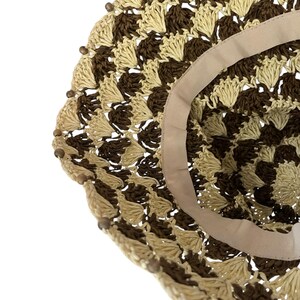 May include: A close-up of a woven sun hat with a brown and beige pattern. The hat has a wide brim and a beige ribbon lining the inside. Small wooden beads are attached around the edge of the brim.