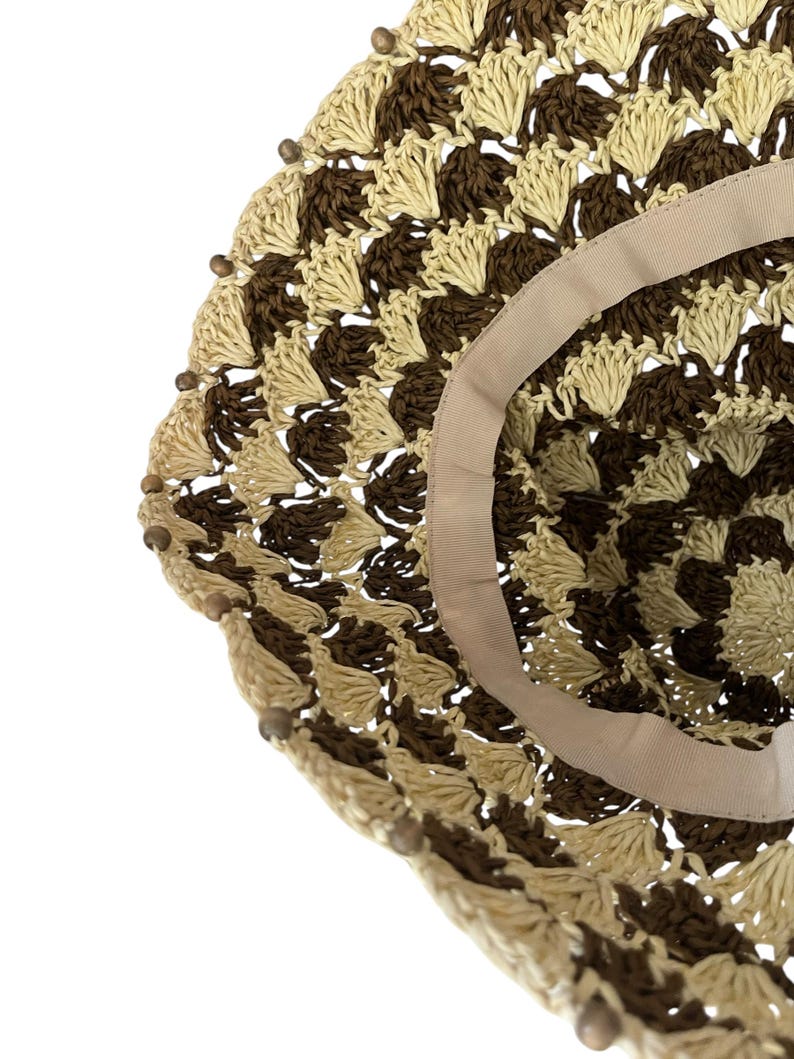 May include: A close-up of a woven sun hat with a brown and beige pattern. The hat has a wide brim and a beige ribbon lining the inside. Small wooden beads are attached around the edge of the brim.