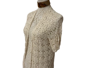 May include: A cream-colored crocheted cardigan with a delicate openwork pattern. The cardigan has a V-neckline and short sleeves.