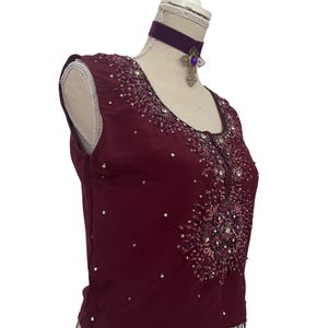 May include: Burgundy sleeveless top with intricate sequin and bead embellishments. The top features a beaded fringe along the bottom hem and a decorative design on the front. A purple velvet choker with a cross pendant is also visible.