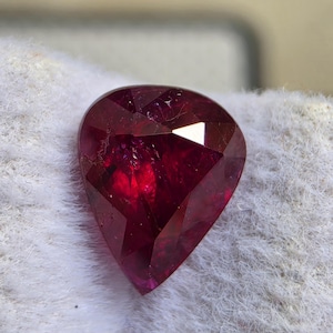 Può includere: Una gemma sfaccettata a forma di pera di colore rosso intenso e saturo. La pietra presenta molteplici sfaccettature triangolari e trapezoidali che riflettono la luce, creando un aspetto brillante e traslucido con inclusioni interne visibili.