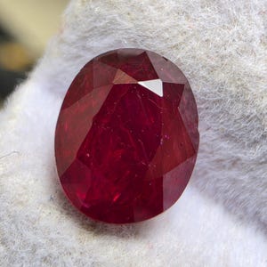 Puede incluir: Una piedra preciosa de color rojo intenso con corte ovalado y una superficie facetada que refleja la luz. La piedra muestra un tono carmesí rico y saturado con inclusiones internas visibles y facetas geométricas nítidas y pulidas.