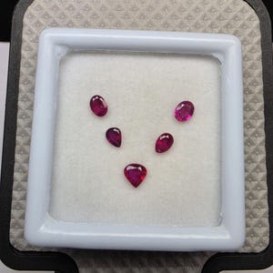 Puede incluir: Cinco piedras preciosas facetadas de tono rojo magenta intenso, dispuestas en forma de V sobre una base de espuma blanca dentro de un estuche cuadrado. La colección incluye dos piedras ovaladas, dos en forma de pera y una más grande.