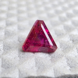 May include: A triangular-cut gemstone with a deep, vibrant magenta-red color. The stone features multiple faceted surfaces that reflect light, creating internal depth and visible inclusions within the translucent, crystalline structure of the gem.